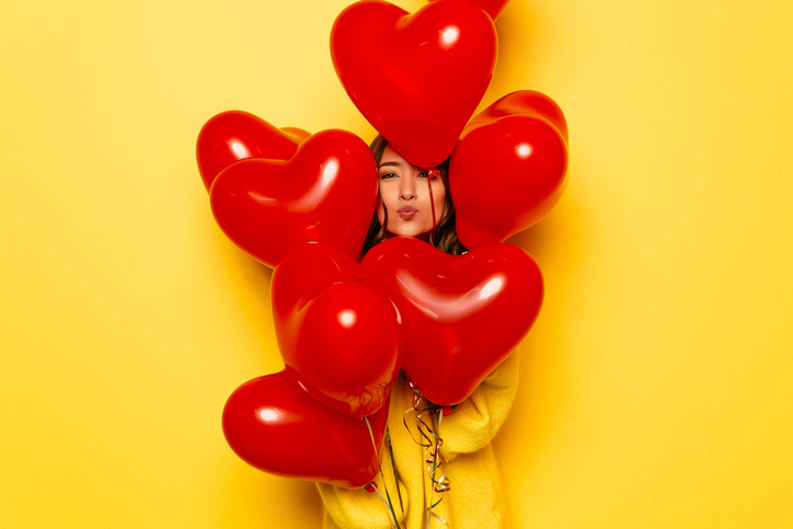 Woman's face hiding between red balloons on yellow background.